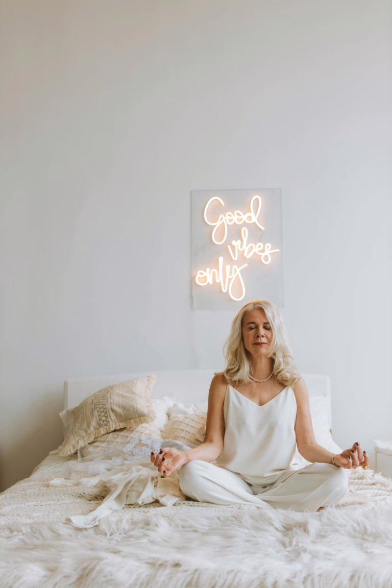 Senior woman meditating on bed with 'Good vibes only' neon sign, embracing tranquility.