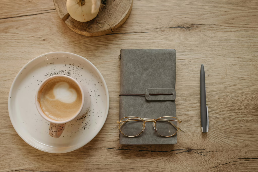 Top view of a cappuccino, notebook, eyeglasses, and pen on a wooden table for a cozy workspace atmosphere.