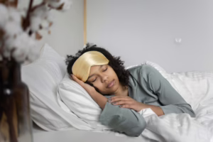 A young woman peacefully sleeps with a sleep mask, nestled in cozy white bedding.